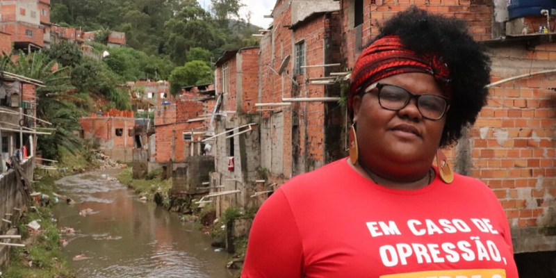 Mulher está em pé em um lugar a céu aberto na Brasilândia. Atrás dela há casas e um rio.