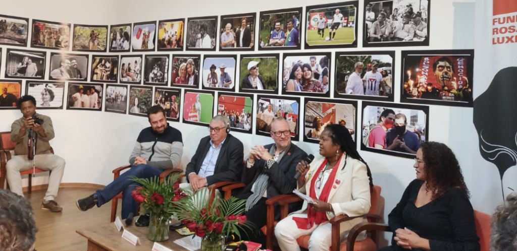 Na foto, mulher negra fala ao microfone, ao lado de outras quatro pessoas, na parede ao fundo há uma exposição de fotos