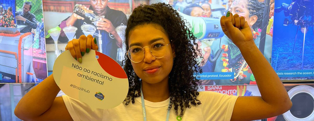 Foto mostra Amanda Costa na COP 27 segurando uma placa com o texto Não ao racismo ambiental