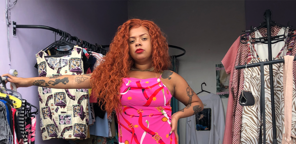 Foto mostra Larissa com uma lace ruiva. Ao fundo vê-se as roupas do Brechó do Barulho