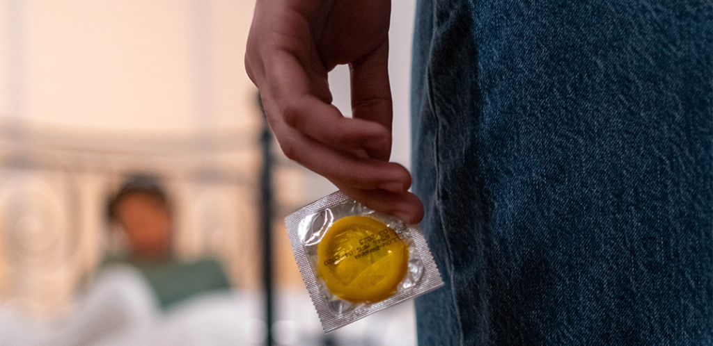 Foto mostra, em primeiro plano, mulher segurando camisinha. Ao fundo, homem na cama