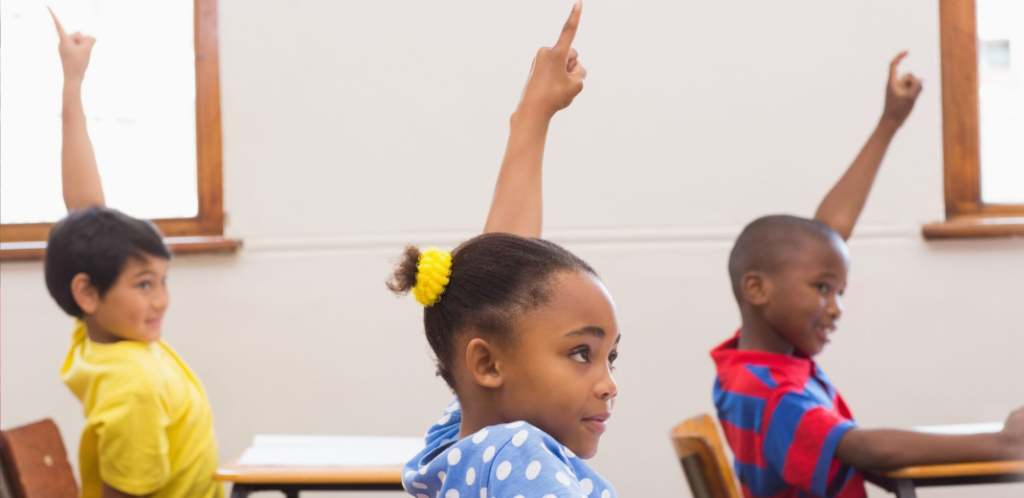 Foto mostra três crianças com as mãos levantadas na escola