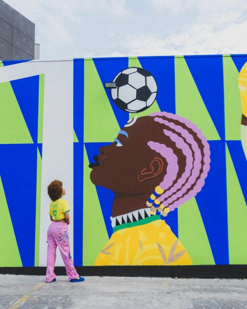 Foto mostra uma a grafiteira Criola, e ao fundo sua obra