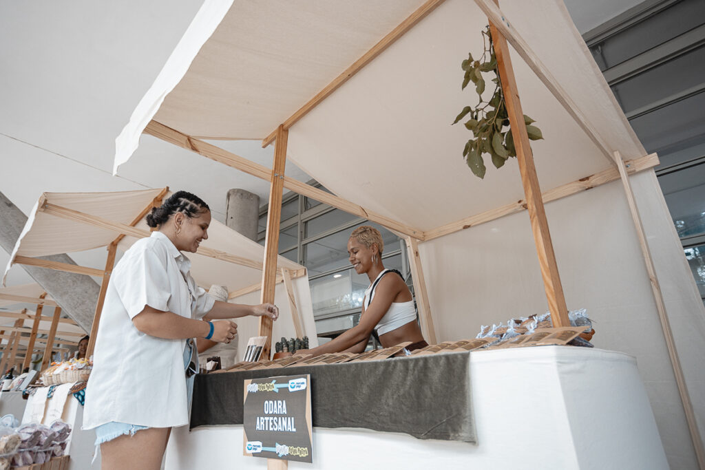 Uma banca de feira, em que uma mulher preta de pele clara com cabelos curtos e platinados exibe seus produtos artesanais para outra mulher preta de pele clara, vestida com camisa branca e shorts