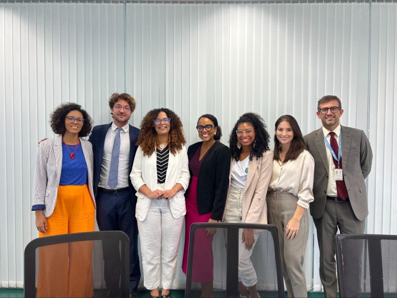 Quatro mulheres pretas e uma mulher branca lado a lado com dois homens brancos de terno e gravata, todos vestidos socialmente. Ao fundo, persianas brancas e na frente, três cadeiras de escritório pretas