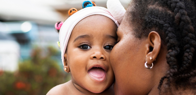 Foto mostra criança negra na primeira infância sorrindo no colo da mãe