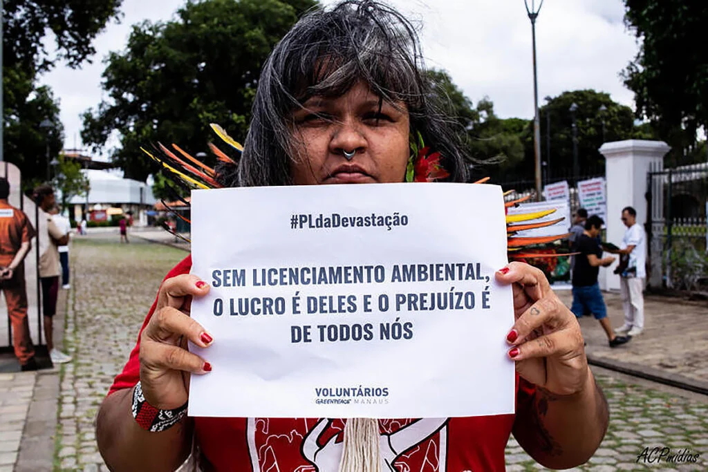 Foto mostra mulher indígena com cartaz contra o racismo ambiental e o PL da Devastação