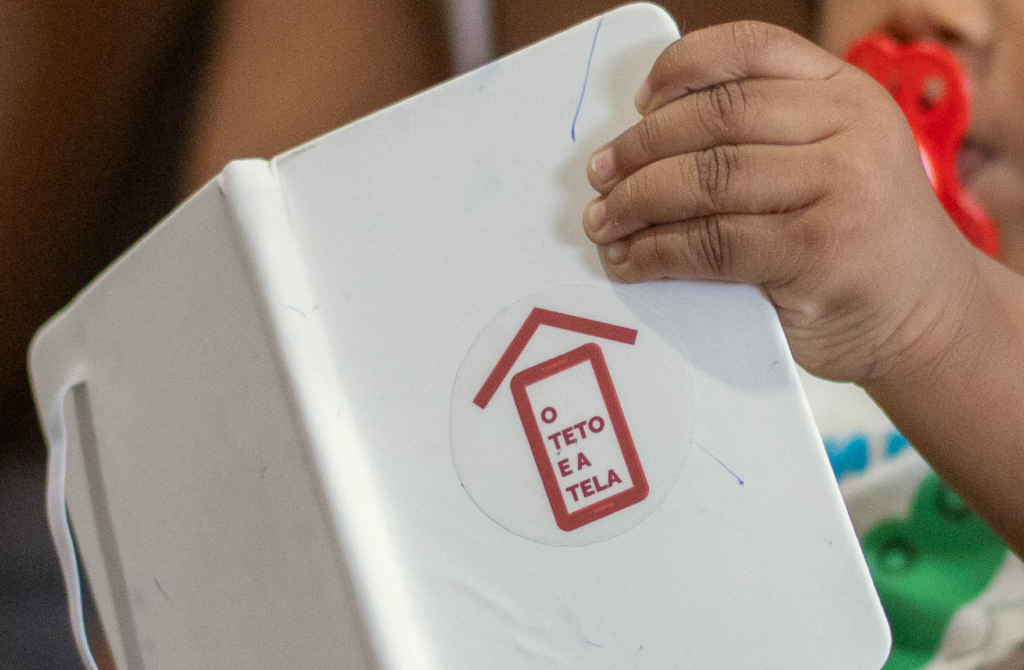Foto mostra criança segurando caderno com o logo do projeto "O Teto e a Tela", que investigou a infância digital nas periferias