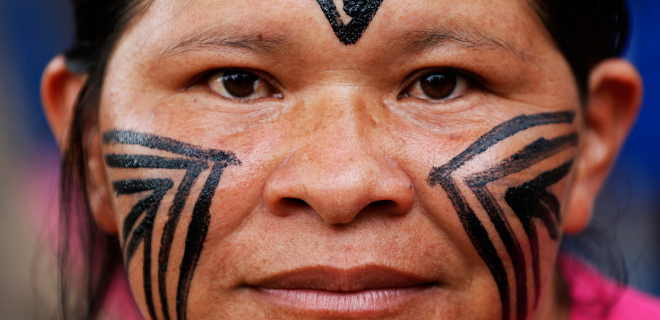 Foto representando as mulheres indígenas brasileiras mostra mulher indígena em close, olhando diretamente para a câmera, com pinturas tradicionais pretas no rosto — linhas geométricas nas bochechas e um desenho central na testa.