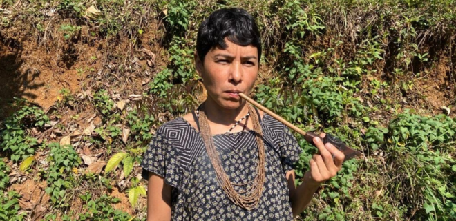 Mulher indígena de cabelo curto segura um cachimbo, em pé em área de mata. Ela usa vestido estampado e colares de sementes, com vegetação densa ao redor.